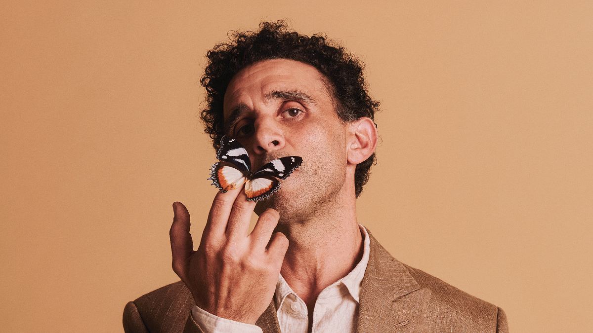 Promotional image for Coriolanus by Bell Shakespeare: a man looks down the camera while holding a butterfly to his lips