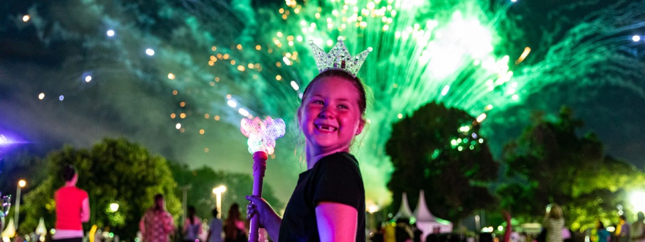 A young girl smiles while carrying a light up wand while a burst of green fireworks explodes in the sky behind her