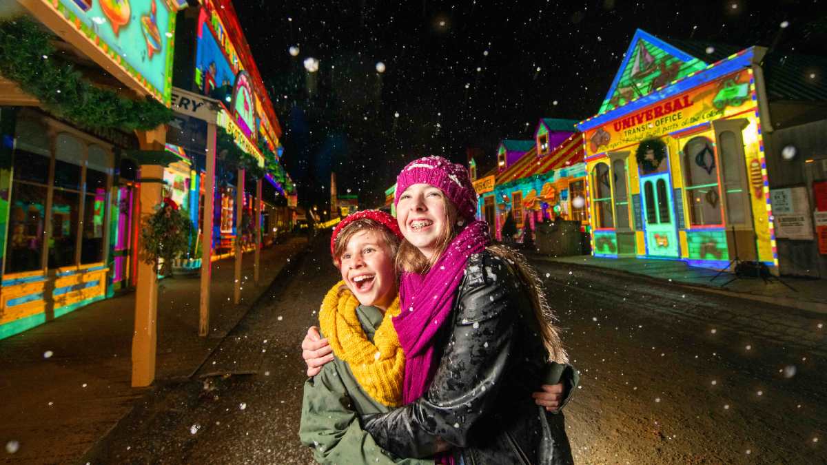 young boy and girl embracing under snow with lit-up historic buildings in background
