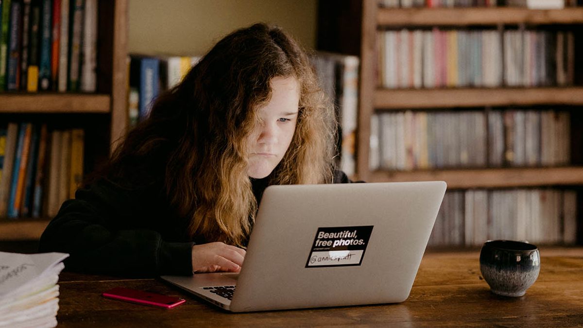 child with long hair working on a laptop at a wooden table with books behind her