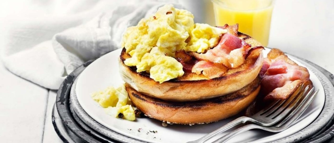 Scrambled eggs and bacon on two muffins on a white plate with two forks.