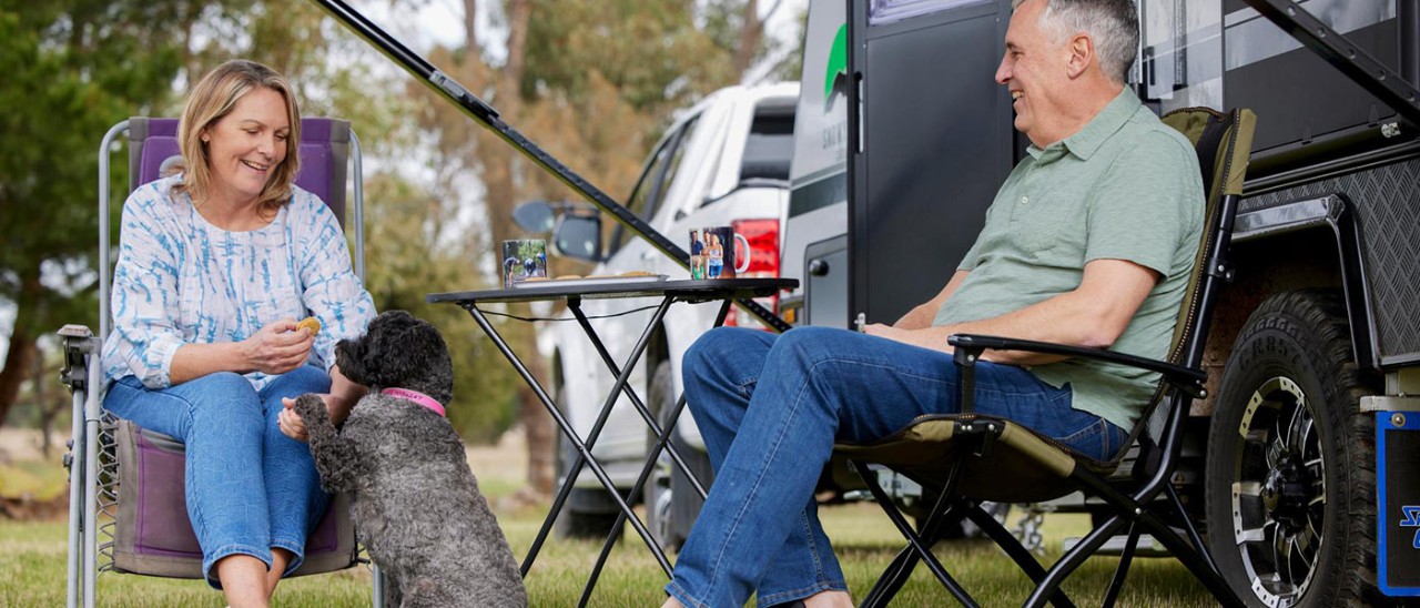 An older couple sitting on deck chairs in front of their parked caravan with their black spoodle dog