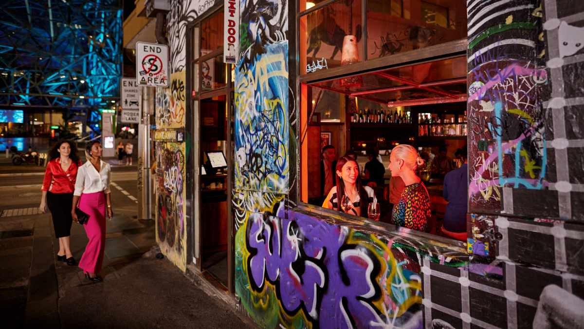 two women walking towards a restaurant covered in graffiti; two women sit in its window