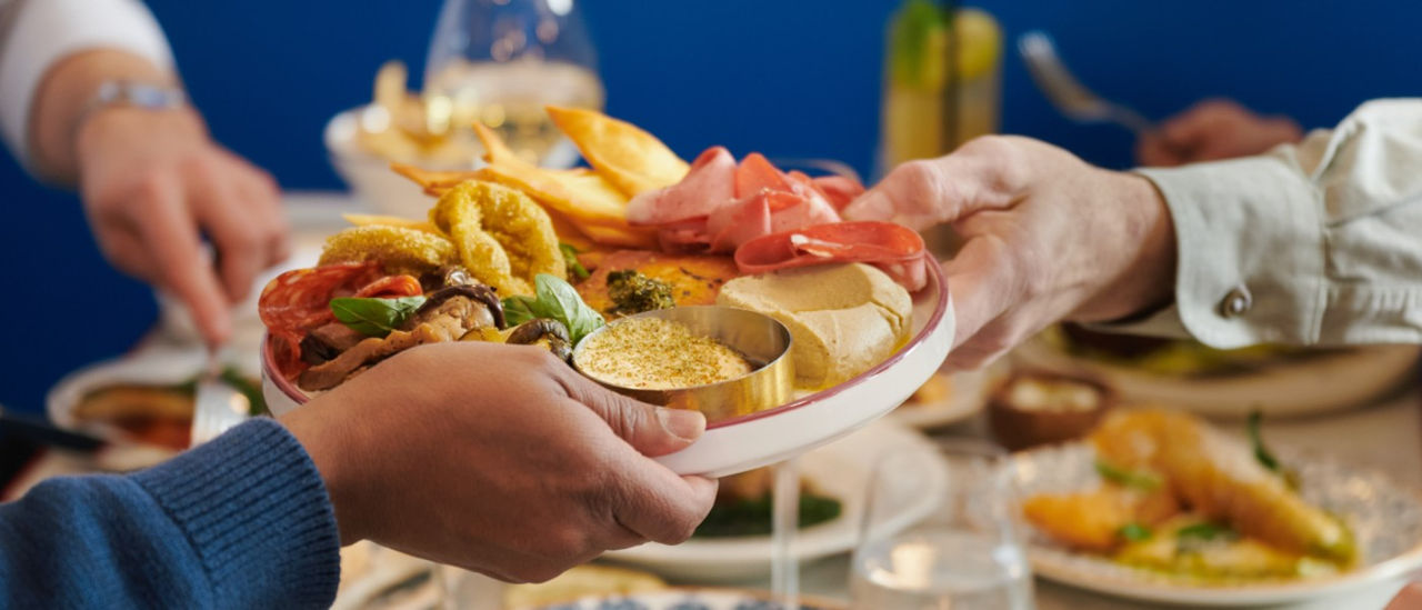 Two people passing a Greek-inspired mezze plate at Sojourn restaurant as part of a larger group meal