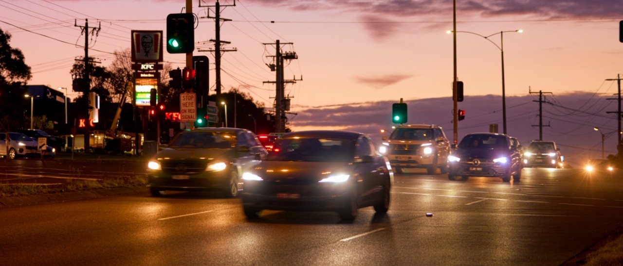 Several cars driving through the intersection of Wantirna and Boronia roads in Wantirna. It is early evening and darkness nearly fallen and the cars have their headlights on