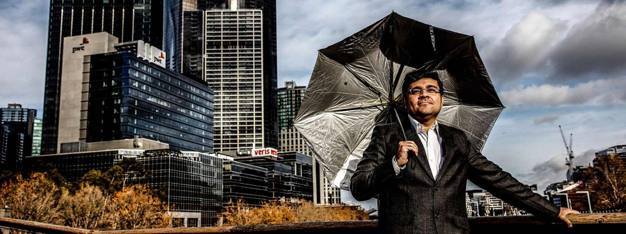 Man with umbrella stands outside with buildings and grey sky in background.
