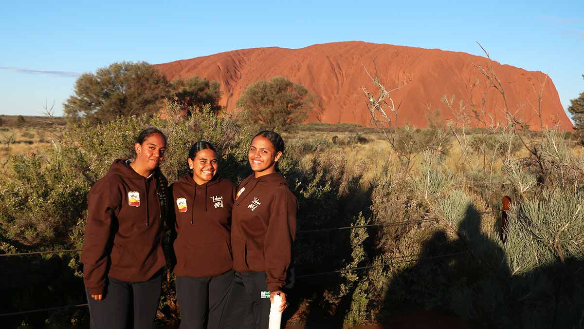 Three school students smiling at camera with Uluru behind.
