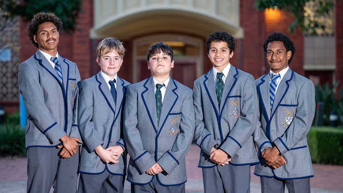 Five teenage boys in school uniform smiling at camera with brick building behind.