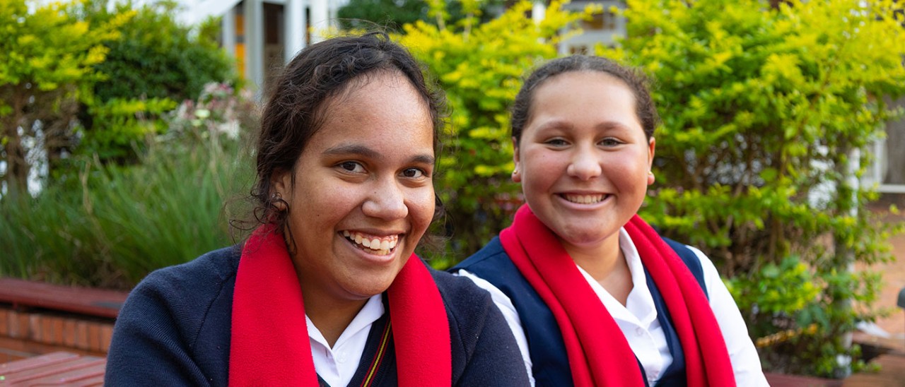 Yalari-15-1400x600.jpg Two school students smiling at camera with garden behind.