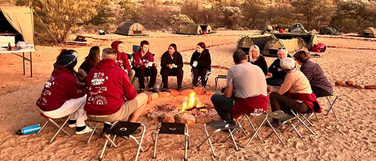 Thirteen people sit around a camp fire at a Yalari Outback Camp in Australia.