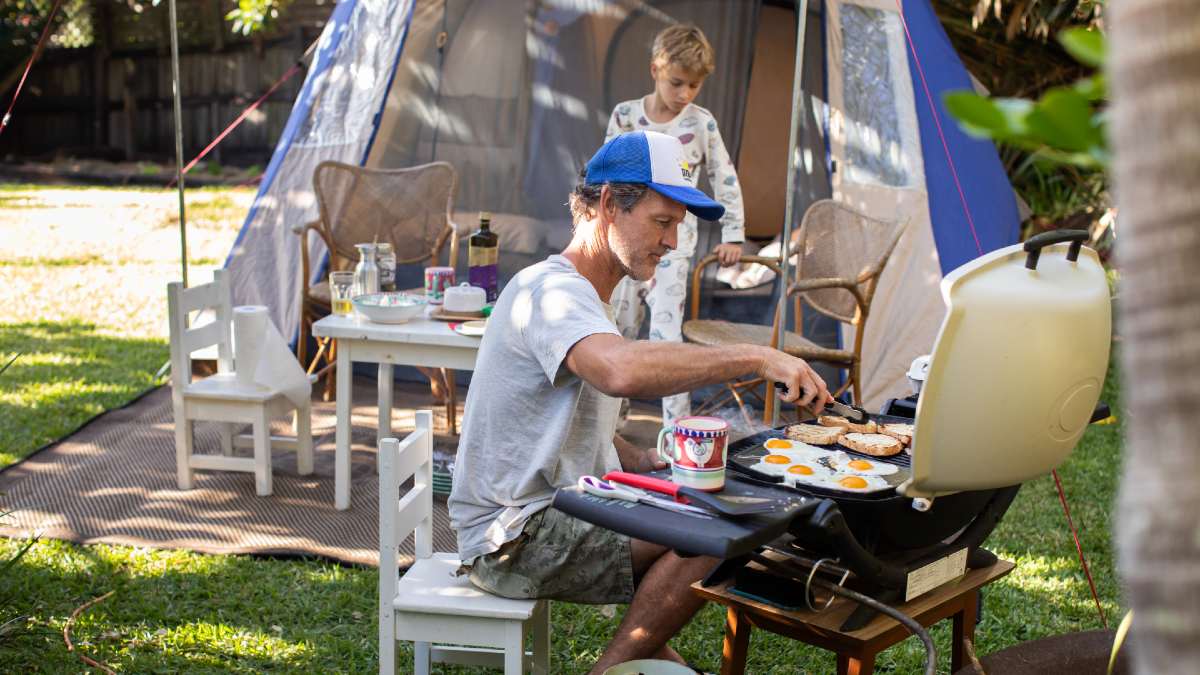 father cooks up breakfast on a camp BBQ outside a tent in front of his son