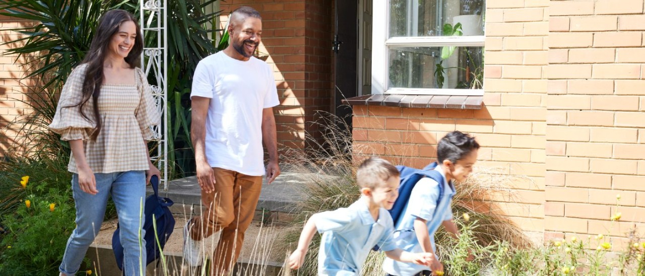 A young family - a man, a woman and two young children in school uniform, smile as they walk outside their brick house on a sunny day