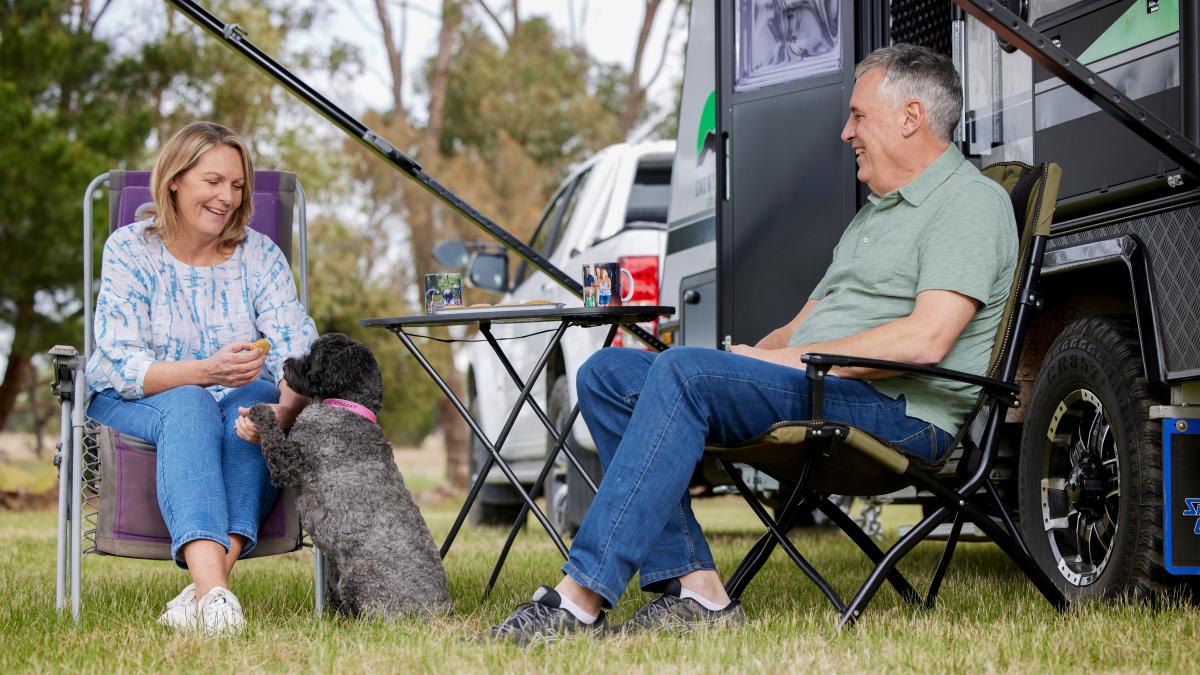 A senior man and woman sitting outside their caravan at a campsite. They are smiling as their black, curly-coated dog begs the woman for a treat