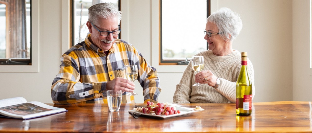 A senior man and woman smiling as they enjoy wine and a fruit platter at home