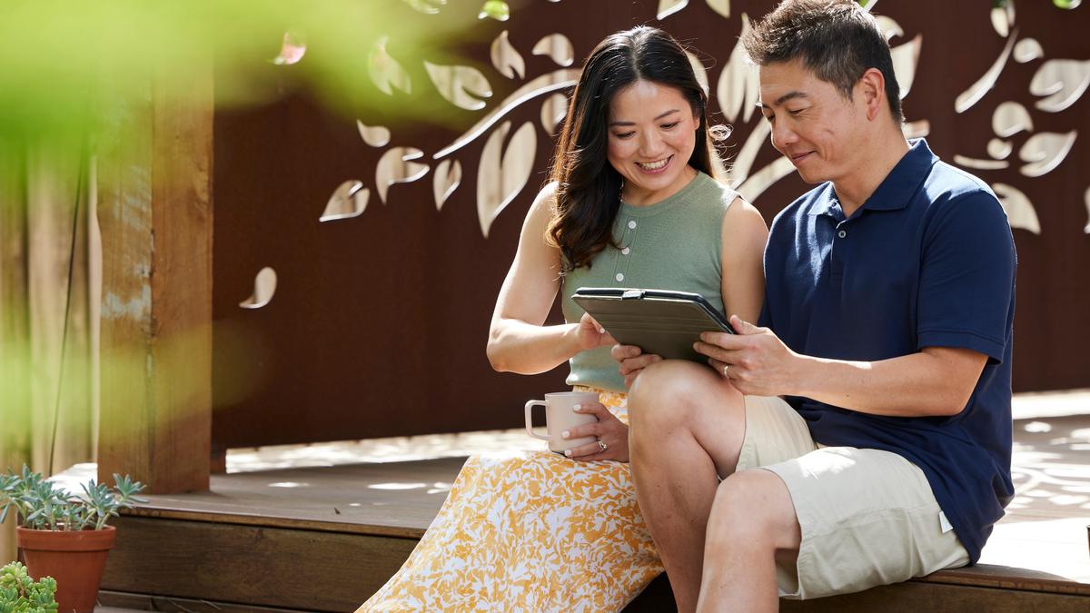 A man and a woman smile happily at a tablet the man is holding. They are both sitting on a wooden deck outside with a oxidised metal wall decal behind them