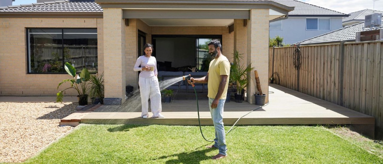 A couple stand in the backyard of a new modern house. The man smiles as he waters the lawn and the woman smiles as she watches him from the deck. It is a sunny day.