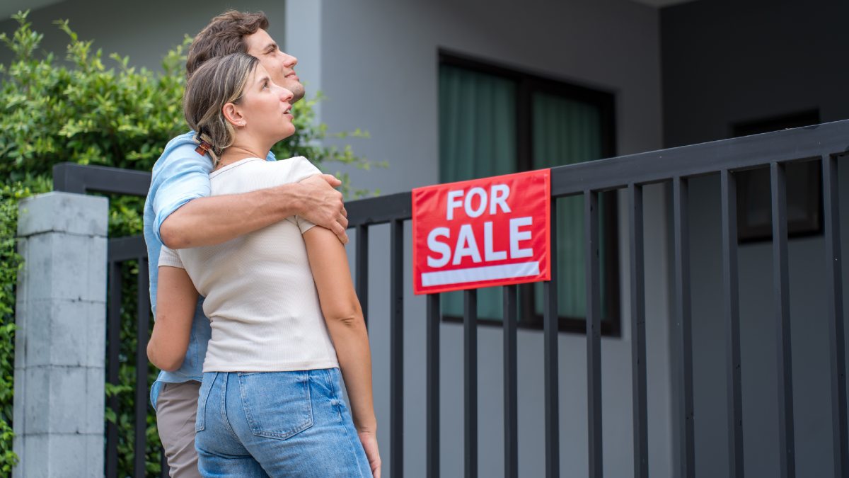 young couple hugging and looking at a home with a for sale sign