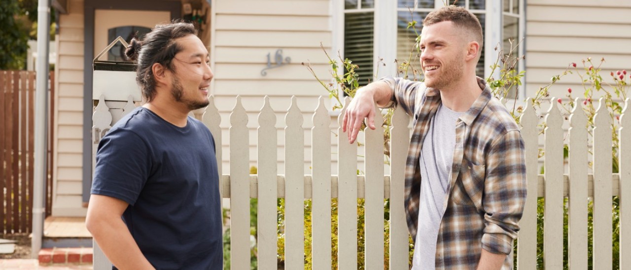 Two men leaning on a picket fence outside a weatherboard house. 