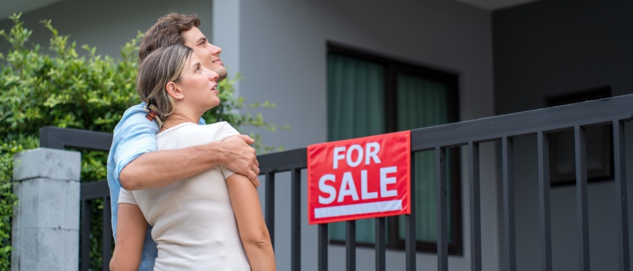 young couple hugging and looking at a home with a for sale sign