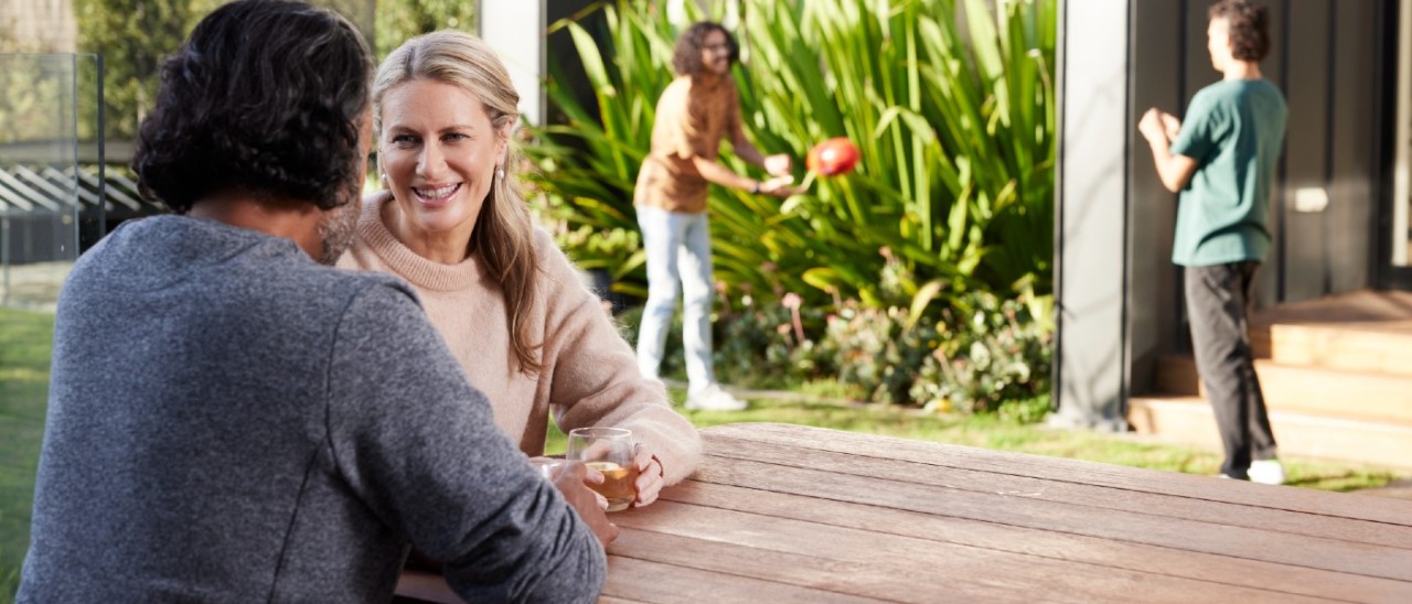 A couple chat happily at a wooden table in their backyard while their two grown children toss a footy to each other in the background