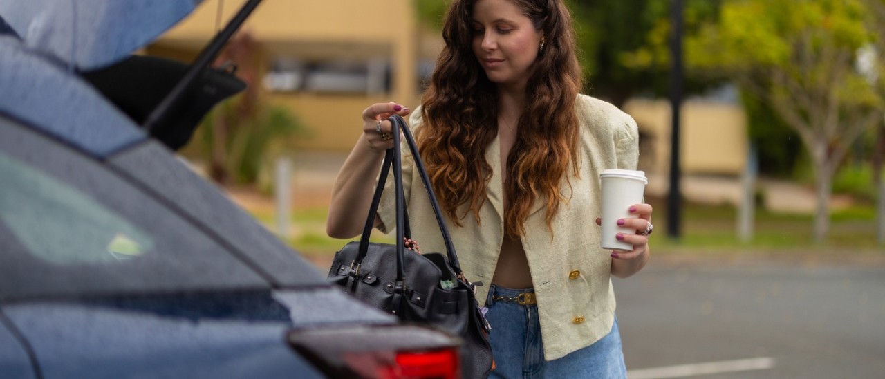 A young woman taking a stylish black leather handbag out of the boot of her car. She is holding a takeaway coffee in her other hand