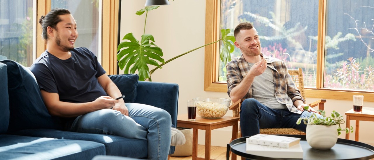 two young men in their living room smiling and watching an offscreen TV