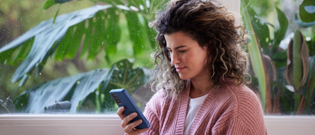 young woman smiling slightly at her smartphone as she sits near a window