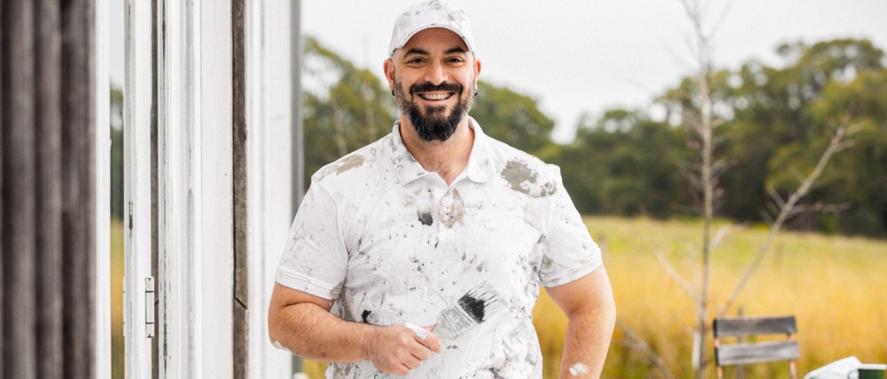 RACV Trades Painter man with paint-covered clothes holding a paintbrush and smiling outside a house