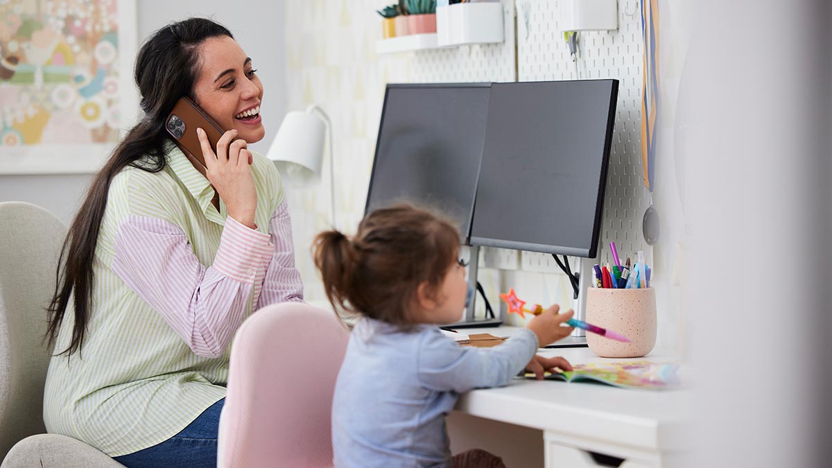 Mother sitting in front of computer at home talking on phone with young daughter playing at desk next to her