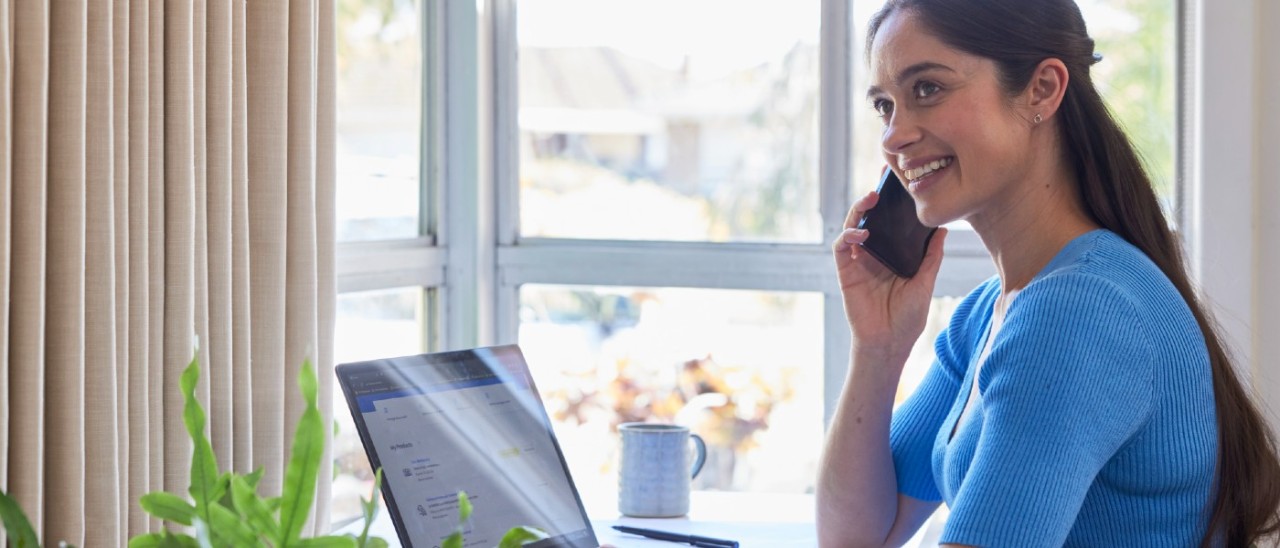 Young woman with long hair and a blue shirt smiling as she speaks on the phone to someone while also using a laptop
