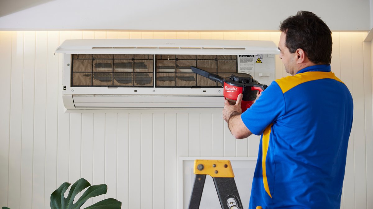 A technician wearing a blue and yellow polo shirt uses a hand-held vacuum to clean an air conditioner filter