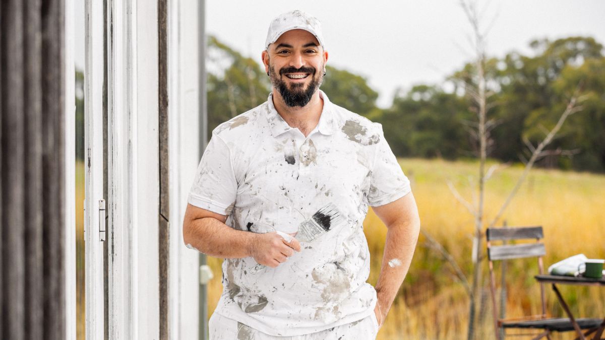 man with paint-covered clothes holding a paintbrush and smiling outside a house