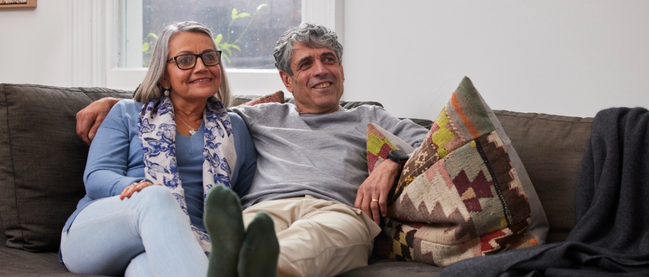 An older couple, a man and a woman, sitting comfortably together on a plush couch. The man rests his feet on a coffee table