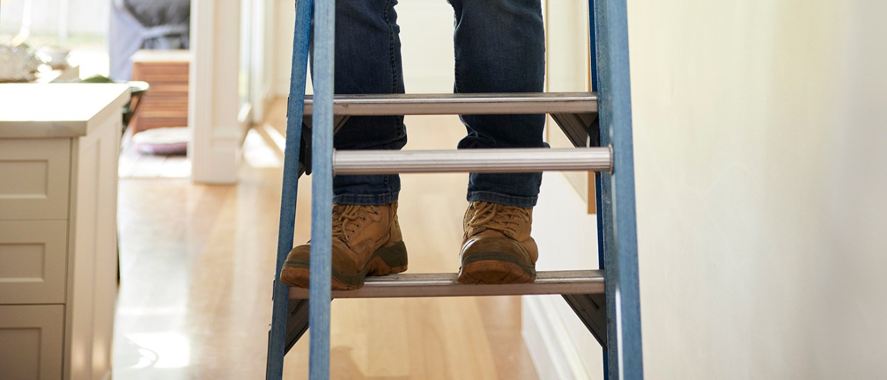 RACV-Trades-ladder-safety-7045-banner-1400x600 Person in jeans with brown boots on a stepladder in hallway with image showing feet on first step