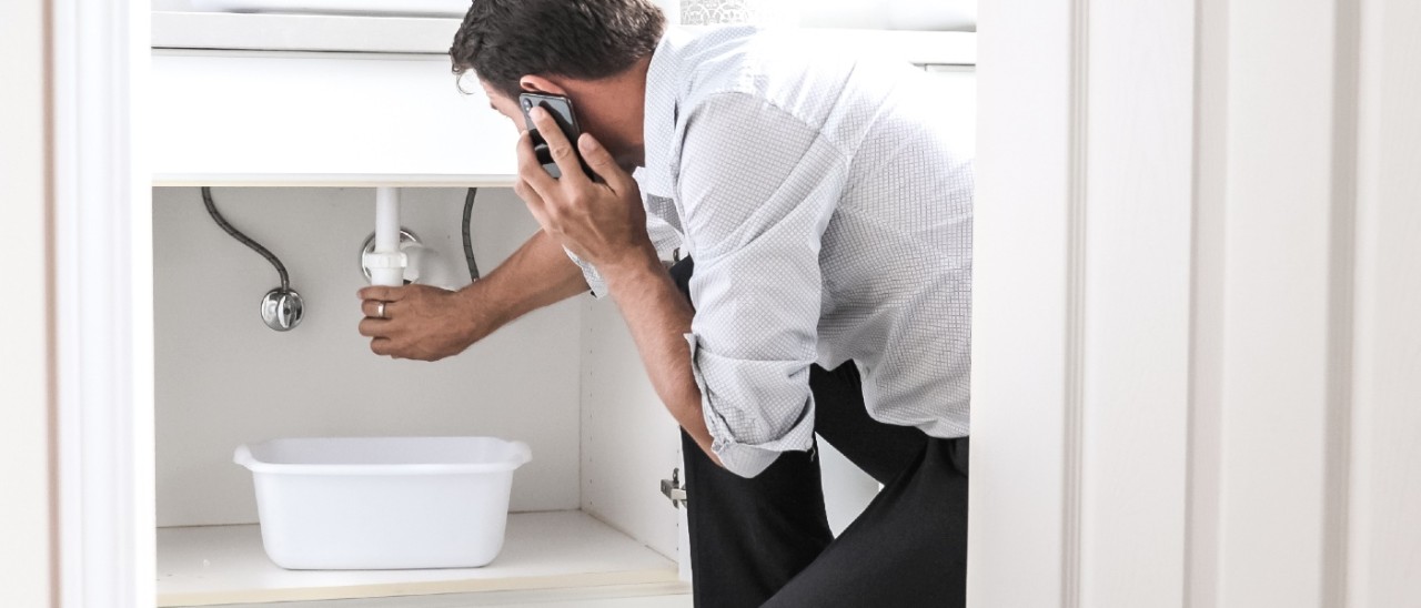 A man calling a plumber as he checks a leaking pipe under a sink