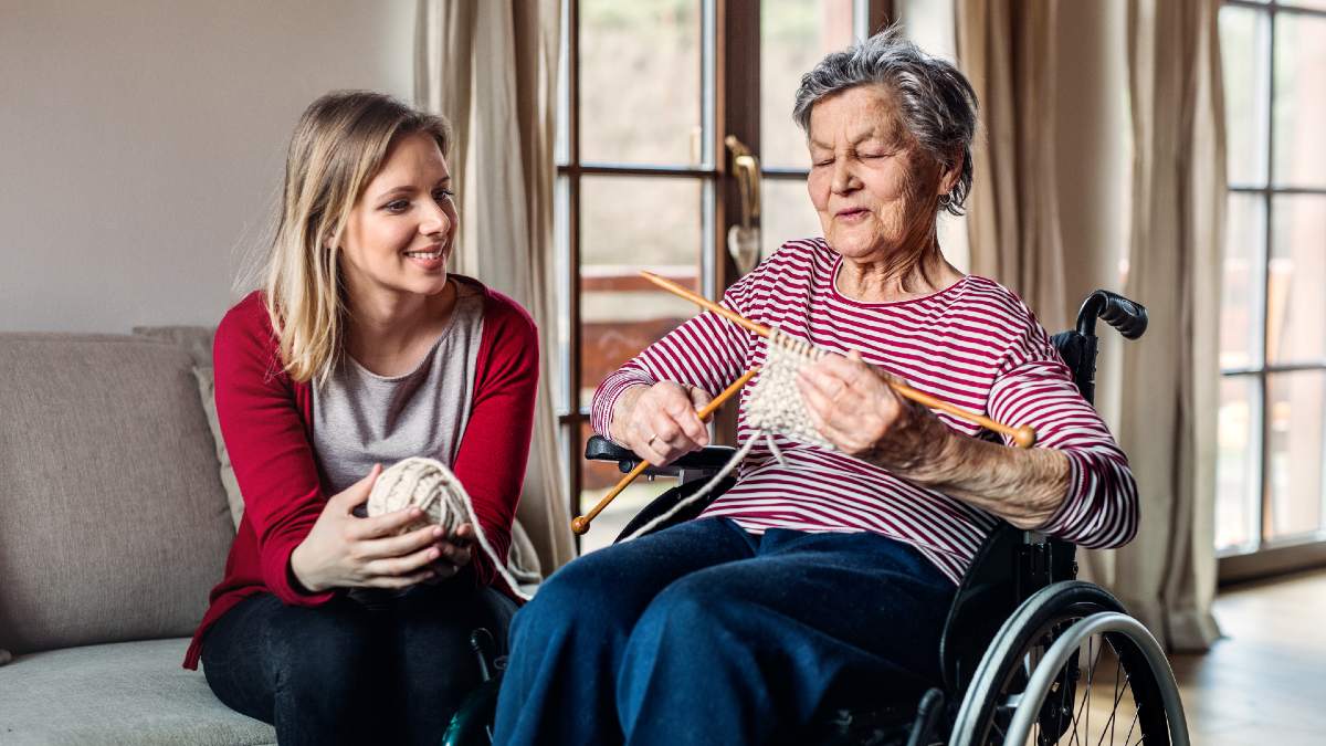senior woman knitting in wheelchair with younger woman holding her yarn