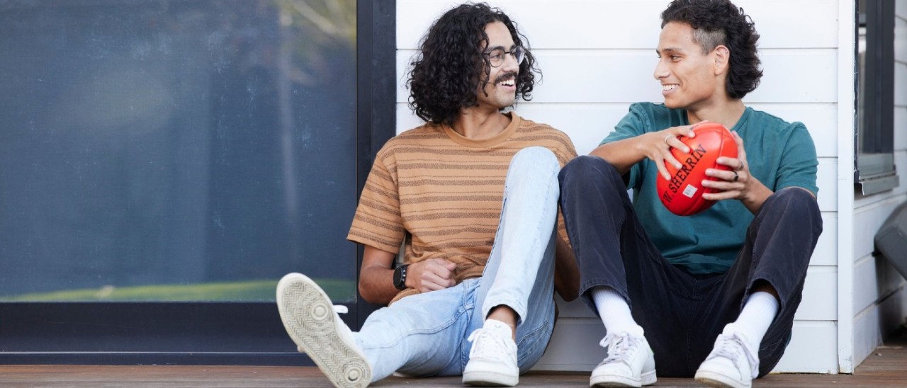 Two young people sitting outside next to a white weatherboard house. They smile at each other and one holds an Aussie Rules  football