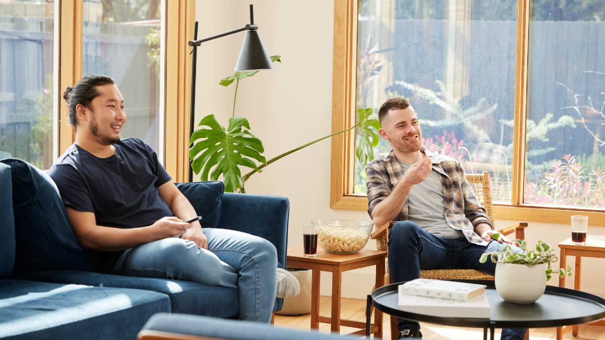 two young men in their living room smiling and watching an offscreen TV