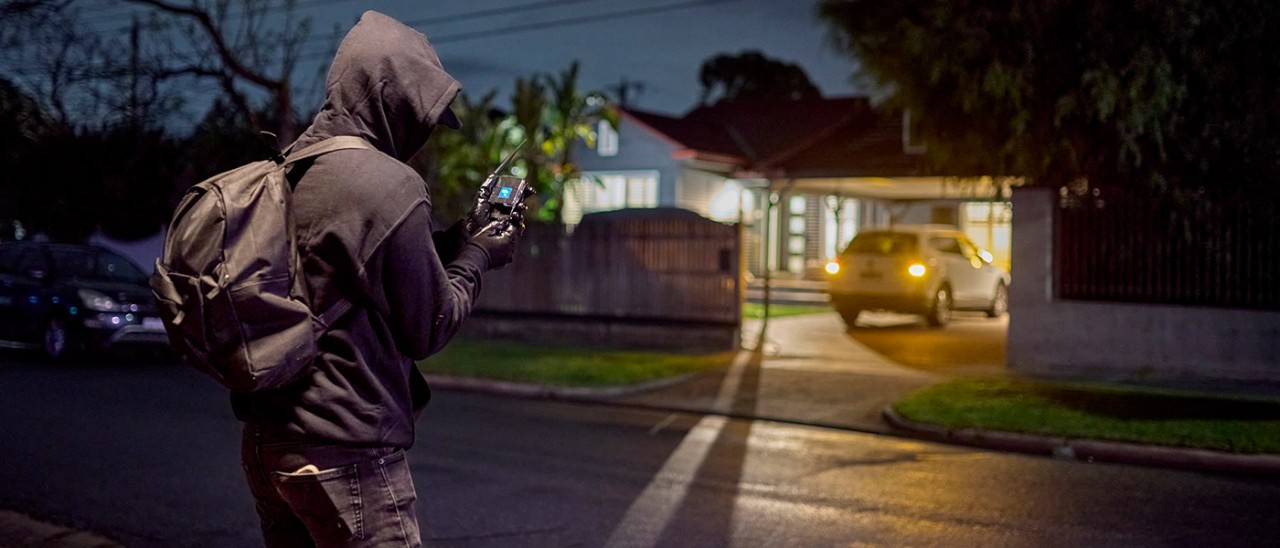 A hooded figure on a street holds a device that appears to be unlocking car in driveway. 