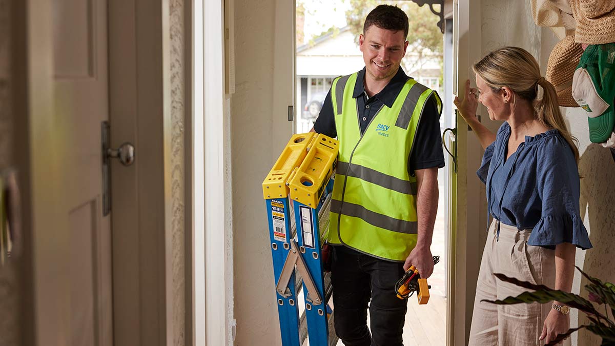 Woman in blue shirt and grey pants opening front door for RACV tradie in hi-vis vest with ladder and tools
