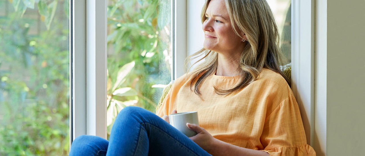 Woman with cup sitting in box window looking at garden. 