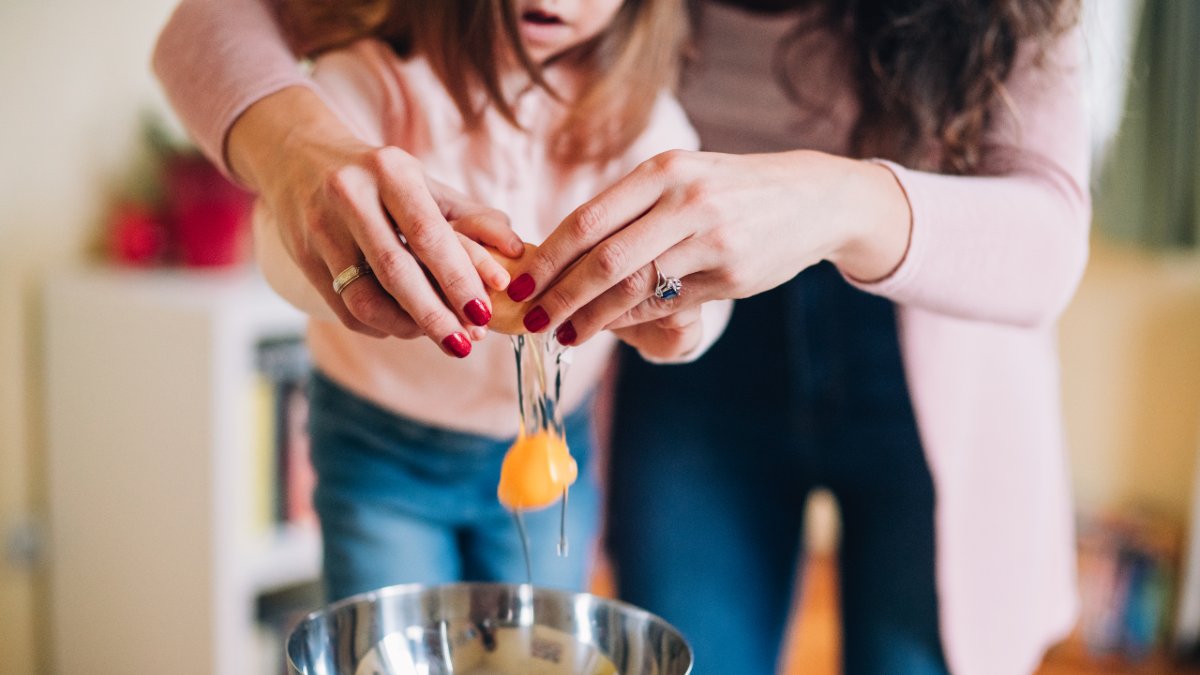 A girl cracking an egg into a frypan with help from her mother.