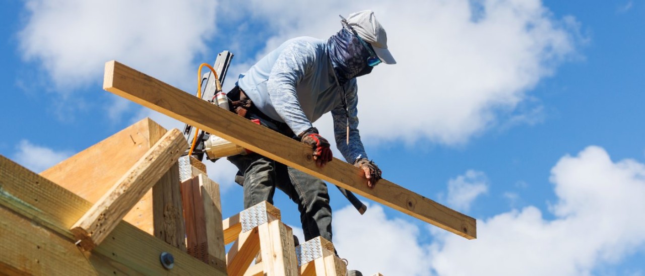 man wearing hat, sunglasses and neck buff carrying a timber plank atop a house construction