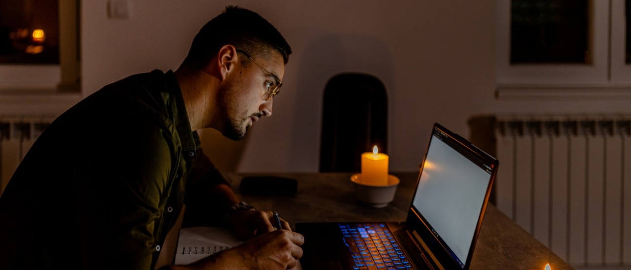 young man taking notes from laptop in a blackout with candles lit nearby