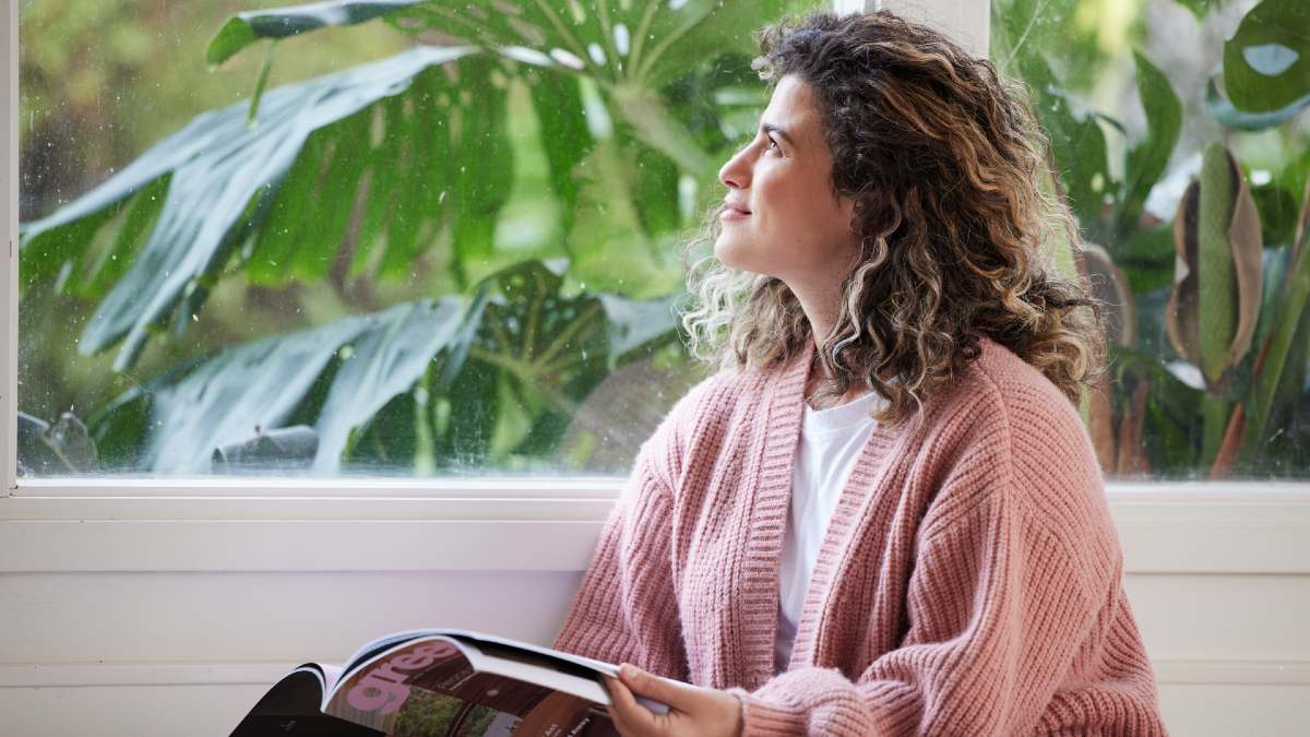 woman in pink cardigan holding a magazine and looking out a window