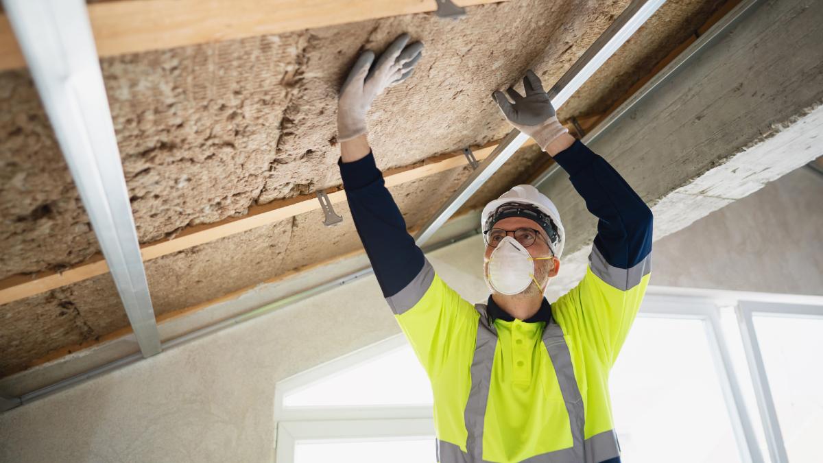 tradie in mask, glasses and hi-vis shirt installing ceiling insulation
