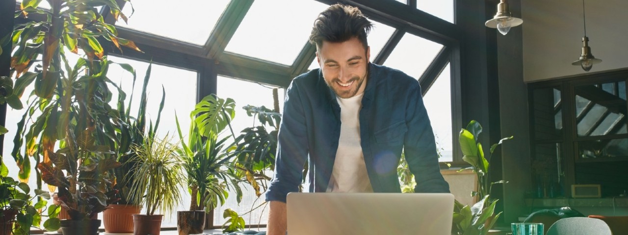 young man smiling at laptop in an office filled with sunlight and green potted plants