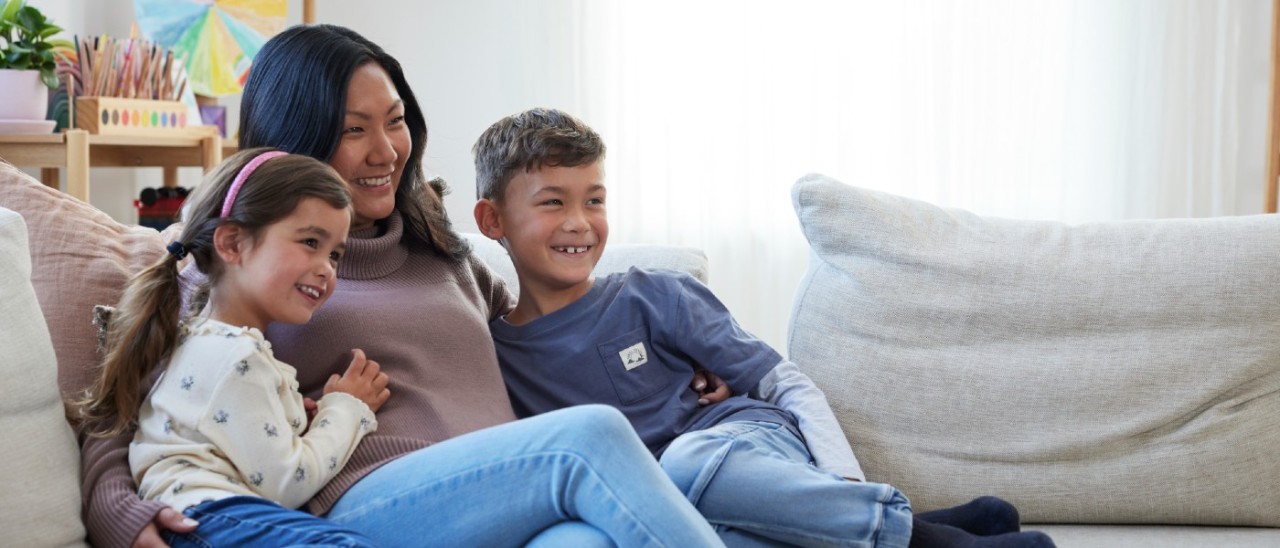 mother, daughter and son cuddling on couch smiling and looking at TV offscreen