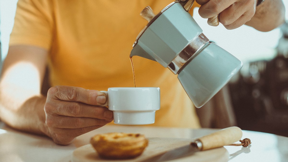 A close-up of a person wearing a yellow t-shirt pouring black coffee into a small white espresso cup from a duck egg blue stovetop coffee maker