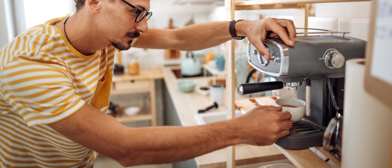 A man wearing glasses and a white and yellow striped t-shirt using a manual espresso machine in a messy home kitchen